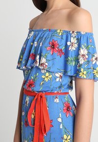 Blaues Off-Shoulder-Kleid mit rot-weißt-gelbem Blumenmuster, Rüschenausschnitt und einem gebundenen roten Gürtel in der Taille.