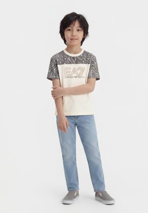 Boy with black hair stands wearing a beige EA7 shirt with patterned sleeves, light blue jeans, and gray slip-on shoes on white background.