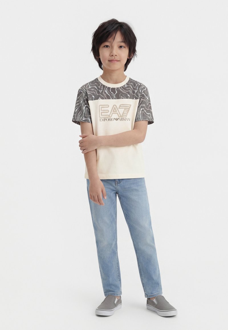 Αγόρι με μαύρα μαλλιά στέκεται φορώντας μια μπεζ μπλούζα EA7 με αμάνικα με σχέδια, ανοιχτόχρωμα μπλε τζιν και γκρι παπούτσια slip-on σε λευκό φόντο.