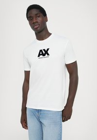 Balta medvilninė marškinė su juodu „AX“ logotipu ir „ARMANI EXCHANGE“ tekstu. Trumpomis rankovėmis ir apvalia iškirpte; standartinė siluetas.