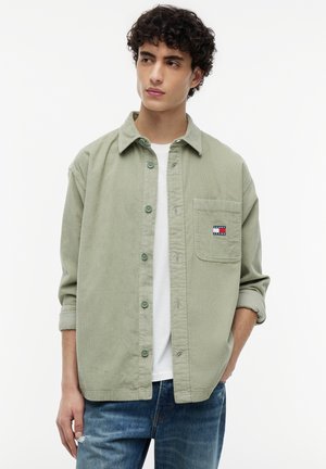 Junger Mann mit lockigem Haar, der ein hellgrünes Hemd mit Knöpfen über einem weißen T-Shirt und blauen Jeans trägt, steht mit einer Hand in der Tasche.