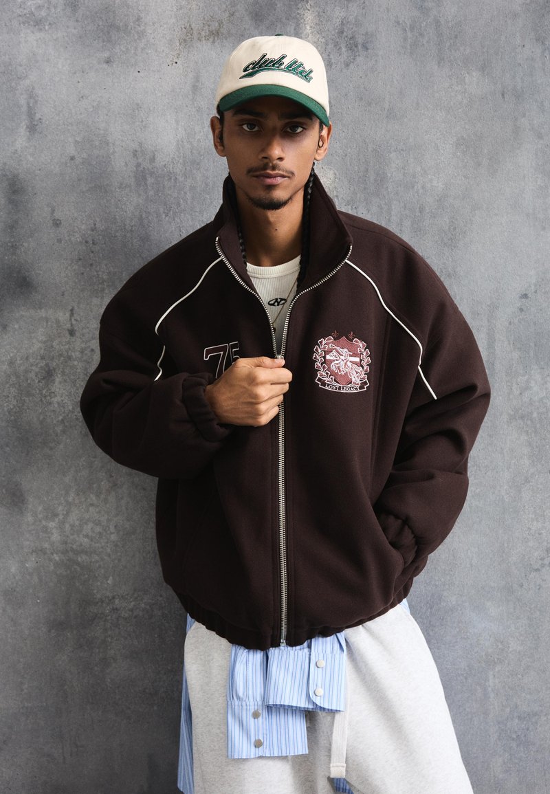 Nuori mies pukeutuneena ruskeaan vetoketjulliseen takkiin, jossa on tunnusmerkki, beige lippalakki, valkoinen paita ja vaaleanharmaat housut, seisomassa harmaata kuvioitua seinää vasten.