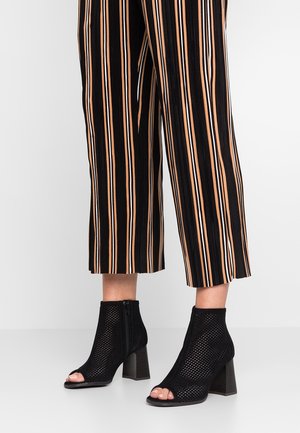 Schwarze offene Knöchelstiefel aus Netzmaterial mit einem dicken Absatz. Kombiniert mit gestreiften, schwarzen und orangefarbenen, cropped Hosen.