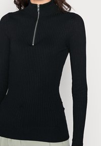Haut noir côtelé à manches longues avec un col montant et une fermeture éclair argentée au niveau du col. Le matériau semble extensible et ajusté, mettant en valeur la silhouette.