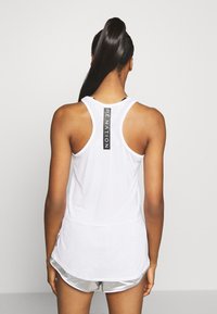 Femme avec de longs cheveux noirs en queue de cheval haute, portant un débardeur blanc avec le texte "Penation" dans le dos et un short de sport argenté, tournée vers le mur.