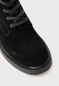 Black suede boot with a rounded toe, features flat gray laces, contrasting stitching, and a black rubber sole for added traction.