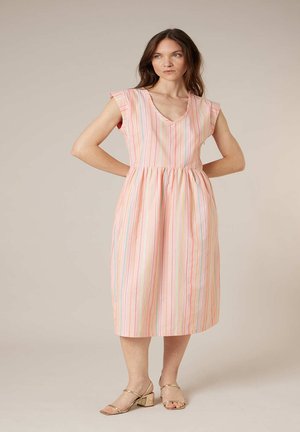 Femme debout, mains sur les hanches, portant une robe midi à rayures roses et des sandales à talons blocs dorés sur un fond uni.