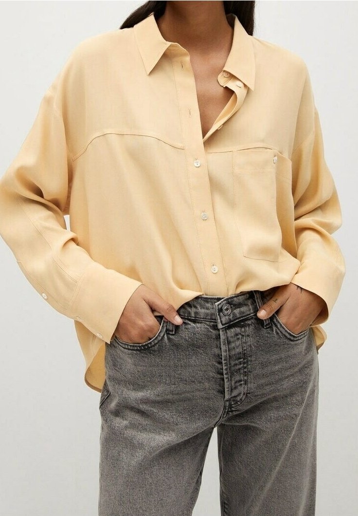 Femme portant une chemise ample à boutons de couleur jaune clair rentrée dans un jean noir délavé taille haute, les mains dans les poches avant.