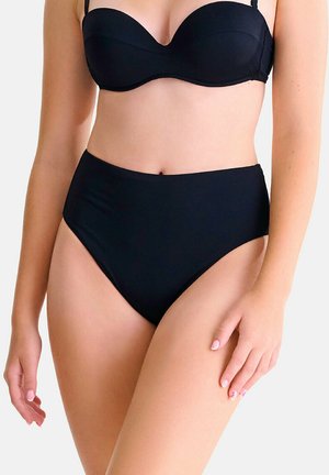 Femme portant un haut de bikini noir sans bretelles et un bas de bikini noir taille haute, debout devant un fond uni.