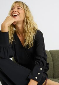 Vêtement noir avec un col en V et des manches bouffantes, orné de boutons en perle aux poignets, porté par une femme aux cheveux blonds ondulés, assise sur un canapé vert.