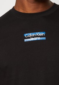 Black cotton T-shirt featuring a blue and white embroidered logo that reads "Calvin Klein Jeans" across the chest in a layered design.