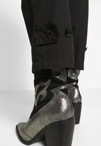 Pantalones negros con bolsillos de botón combinados con botas metálicas plateadas hasta el tobillo que presentan un patrón texturizado y un diseño de tacón ancho.