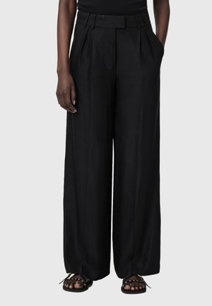 Person som har på seg høytlivs, svarte vidbukser med folder, svarte sandaler med nagler, stående foran en ensfarget lys bakgrunn.