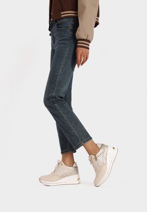 Sneakers beige con tomaia testurizzata, dettagli argentati lucidi e suola ammortizzata. Indossati con jeans blu scuro in una posizione casual.