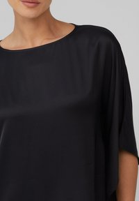 Blouse en soie noire avec un décolleté large et arrondi et des manches dolman courtes. Le tissu a une texture lisse et légèrement brillante.