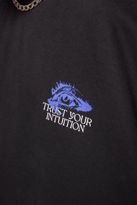 Camiseta de algodón negra con un diseño gráfico de un ojo azul encima del texto "CONFÍA EN TU INTUICIÓN" en fuente serif blanca.