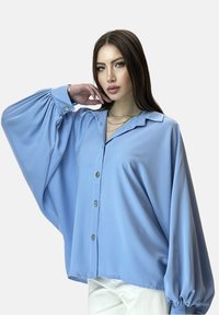 Blusa holgada de color azul claro con mangas largas y voluminosas y cierre de botones en la parte delantera. Tejido suave con un ligero brillo y botones dorados.