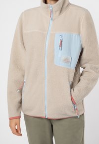 Beige fleecejacka med en ljusblå dragkedjeficka på bröstet, kontrasterande detaljer och hög krage. Har sidodragkedjefickor.