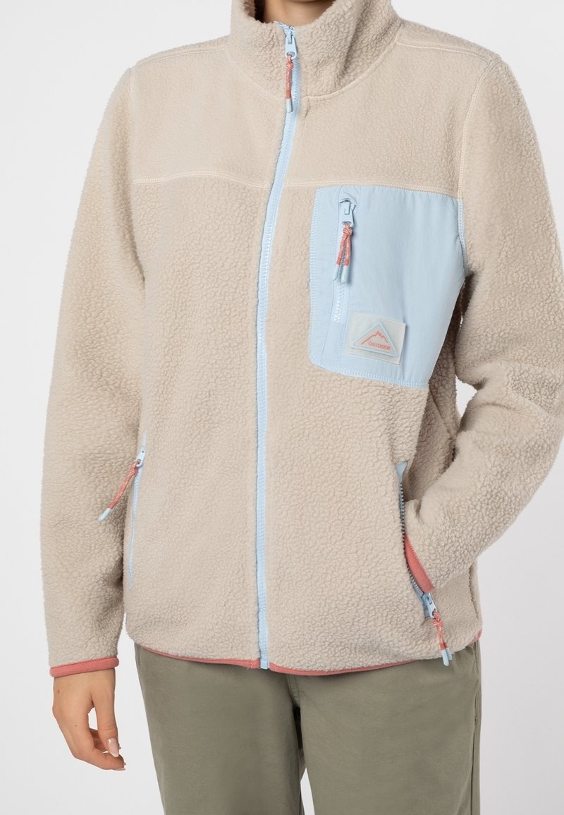 Beige fleecejacka med en ljusblå dragkedjeficka på bröstet, kontrasterande detaljer och hög krage. Har sidodragkedjefickor.