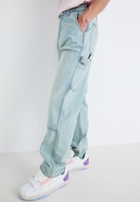 Pantalones cargo de mezclilla de lavado claro con un ajuste relajado, que presentan múltiples bolsillos y una superficie suave, ligeramente texturizada. Combinados con zapatillas blancas.
