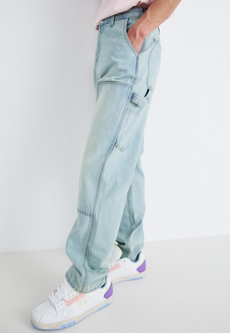 Pantalones cargo de mezclilla de lavado claro con un ajuste relajado, que presentan múltiples bolsillos y una superficie suave, ligeramente texturizada. Combinados con zapatillas blancas.