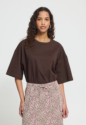 Kvinde med krøllet hår iført en løs mørkebrun T-shirt og et højtsiddende, lyserødt og brunt leopardprint nederdel mod en ensfarvet baggrund.