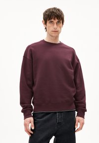 Felpa bordeaux con collo e polsini a costine, presenta una vestibilità ampia e una consistenza morbida. Abbinata a pantaloni in jeans scuri.