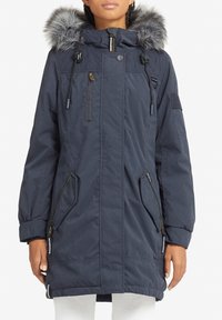 Navy parka with fur-trimmed hood, front zipper, two zippered pockets, and quilted accents. Features a relaxed fit and elastic cuffs.