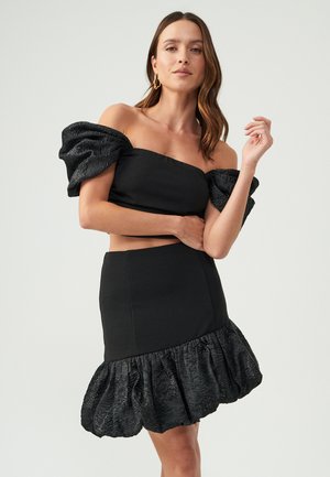 Zwart off-the-shoulder jurkje met poefmouwen, getailleerde buste en een gerimpelde textuur aan de onderkant. Gemaakt van een gladde stof met een subtiele glans.