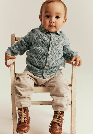 Strikket, lyseblå cardigan med diamantmønstre, knappelukning, parret med grå joggingbukser og brune snørestøvler, siddende på en træstol.