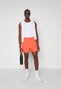 Roupa atlética masculina composta por uma camisola regata branca, calções laranja e ténis brancos, segurando uma garrafa de água preta com óculos de sol azuis.