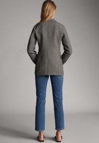 Harmaa räätälöity takki, jossa on lovilaput, tekstuuri ja keskilinjasauma. Yhdistetty sinisiin farkkuihin, joissa on leveät lahkeet, ja mustiin muloihin.