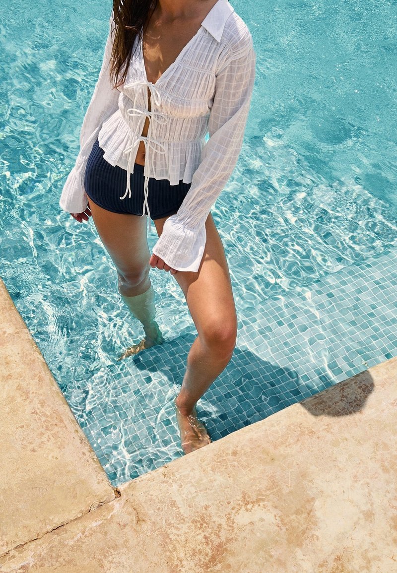 Femme portant un chemisier blanc noué et un short sombre, debout jusqu'aux genoux dans une piscine carrelée près du bord de la piscine.