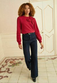 Červený pletený sveter s kvetinovým detailom, skombinovaný s vysokými tmavými džínsovými nohavicami s širokými nohavicami. Model nosí čierne topánky na podpätku. Jednoduché pozadie.