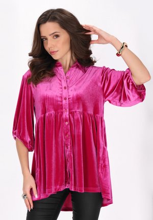 Fuchsia Samtshirt mit Knopfverschluss, kurzen Puffärmeln und einem ausgestellten Saum, kombiniert mit schwarzen Hosen und einem metallischen Armband.