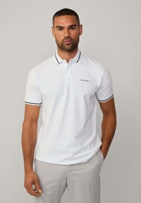 Valkoinen polo-paita mustilla ja laivastonsinisen sävyillä kauluksessa ja hihoissa, jossa on pieni "HACKETT"-logo rinnassa.
