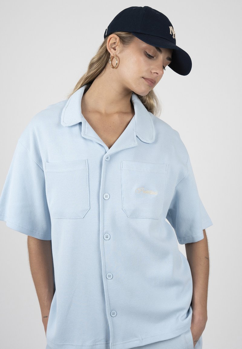 Femme portant une chemise bleu clair à manches courtes boutonnée et une casquette marine, regardant vers le bas, avec des boucles d’oreilles créoles dorées et les mains dans les poches.
