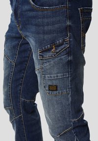 Modré džíny z denimu s více kapsami, kontrastním oranžovým prošitím a látkovou visačkou. Textura je hladká a odolná.