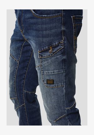 Sinised teksapüksid, millel on mitme taskuga disain, kontrastsed oranžid õmblused ja kangast silt. Materjal tundub sile ja vastupidav.