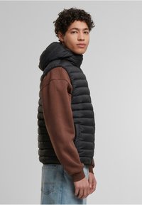 Čierny puffrový vest s kapucňou, s prešívaným dizajnom a rebrovanými manžetami, nosený cez hnedý dlhý rukáv a modré džínsy.