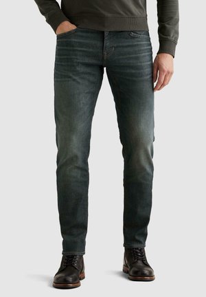 Άνδρας που φοράει σκούρο ξεβάμμενο τζιν slim-fit, μαύρα δερμάτινα μπότες με κορδόνια και σκουροπράσινο μακρυμάνικο τοπ με το ένα χέρι στην τσέπη.