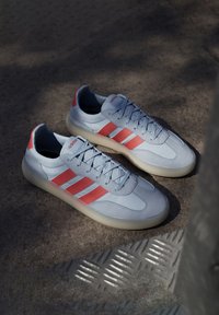 Svijetloplavi tenisice s tri crvene pruge, gumeni potplat, glatka i teksturirana gornja površina, vezanje, logo na jeziku i peti.