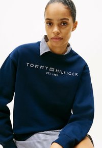 Felpa blu navy con logo bianco "TOMMY HILFIGER EST. 1985", con colletto e polsini a costine, indossata sopra una camicia a righe.