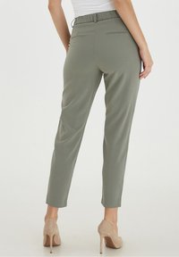 Bakifrån av en person som bär grå slim-fit byxor och beiga högklackade skor, stående mot en enfärgad vit bakgrund.