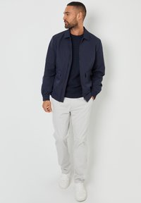 Marineblauer Bomberjacke mit elastischen Bündchen, Reißverschluss und Seitentaschen, kombiniert mit hellgrauen Hosen und weißen Sneakers.