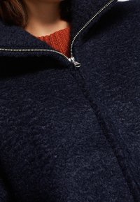 Merinotakki, jossa on korkea kaulus, vetoketjukiinnitys ja teksturoitu kangas. Kauluksen sisäpuolella on kontrastivärinen oranssi neulos.