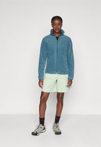 Veste en polaire bleue avec un col haut, une fermeture éclair et un écusson avec logo. Associée à un short vert clair et des chaussures de randonnée grises.