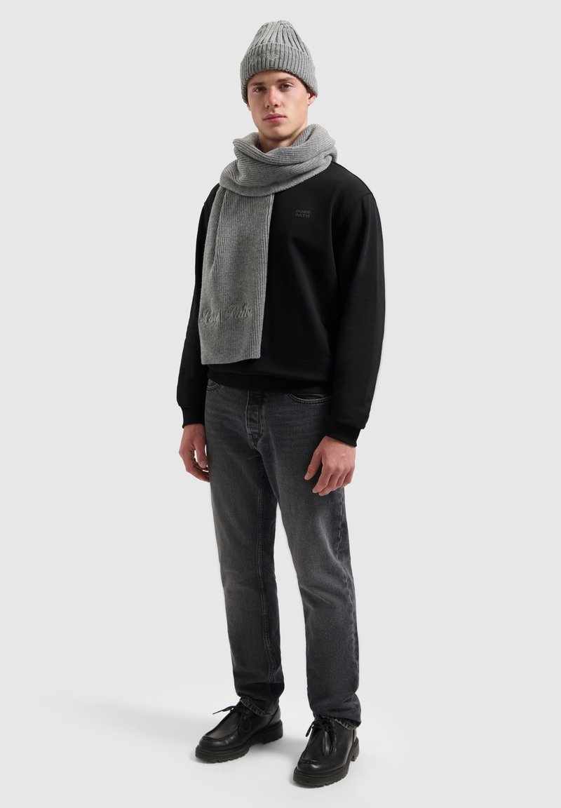 Grijze gebreide beanie en sjaal, zwarte sweatshirt, grijze jeans en zwarte schoenen. De sjaal heeft een geribde textuur en subtiele merkvermelding.