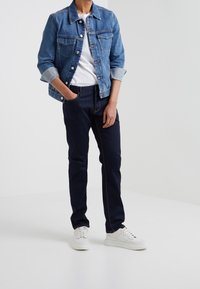 Person klädd i en blå jeansjacka över en vit skjorta, mörkblå jeans och vita sneakers, stående med en hand i fickan.