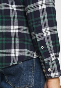 Ruudullinen flanellipaita tummansinisessä, vihreässä, valkoisessa ja harmaassa värissä napillisilla hihansuilla. Kangas on pehmeä. Yhdistetty sinisiin farkkuihin.